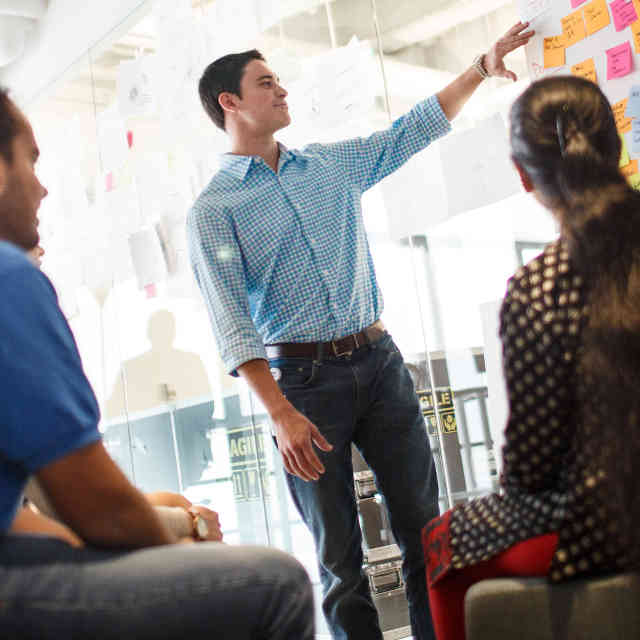 Man gesturing to a board with several post-it notes while two people look on.